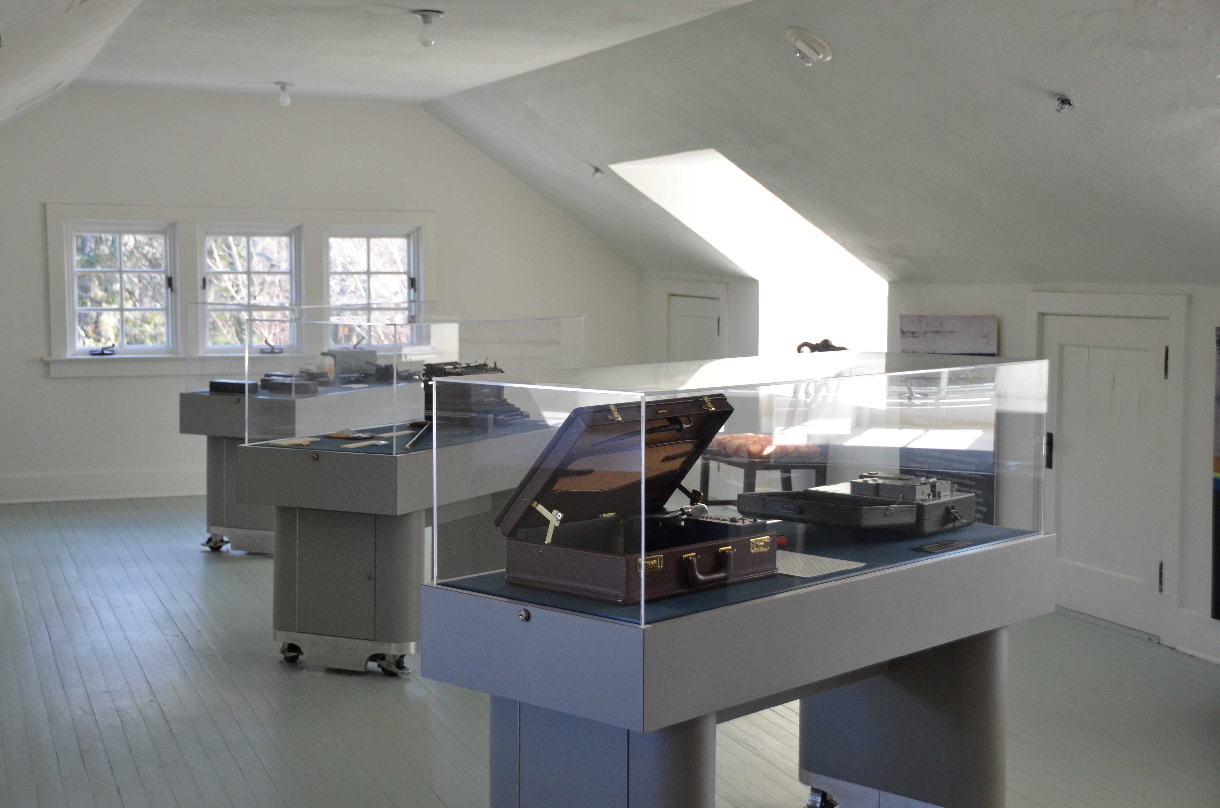 A clean, white-painted attic, lit from sunlight coming through the windows, with three platforms holding enclosed artifacts.